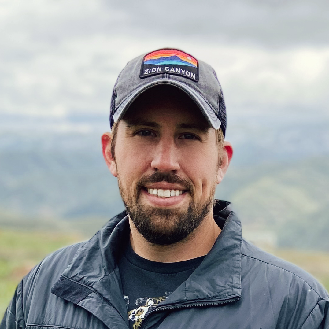 Photo of Andrew Hyte in the mountains, smiling, wearing a zions National Park hat.